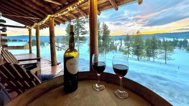 A wooden cabin with a wooden table holding a bottle and two glasses of red wine, overlooking a snowy landscape. - Elk City Escape - Amazing Views, Elk, & Wildlife (Elk City)