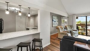 A modern kitchen with a white countertop and bar stools, a dining area with a sofa and chairs, and a large sliding glass door leading to an outdoor patio.