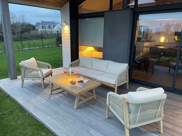 A covered outdoor seating area with a sofa and two chairs, a coffee table with snacks, and a view of a house in the background.