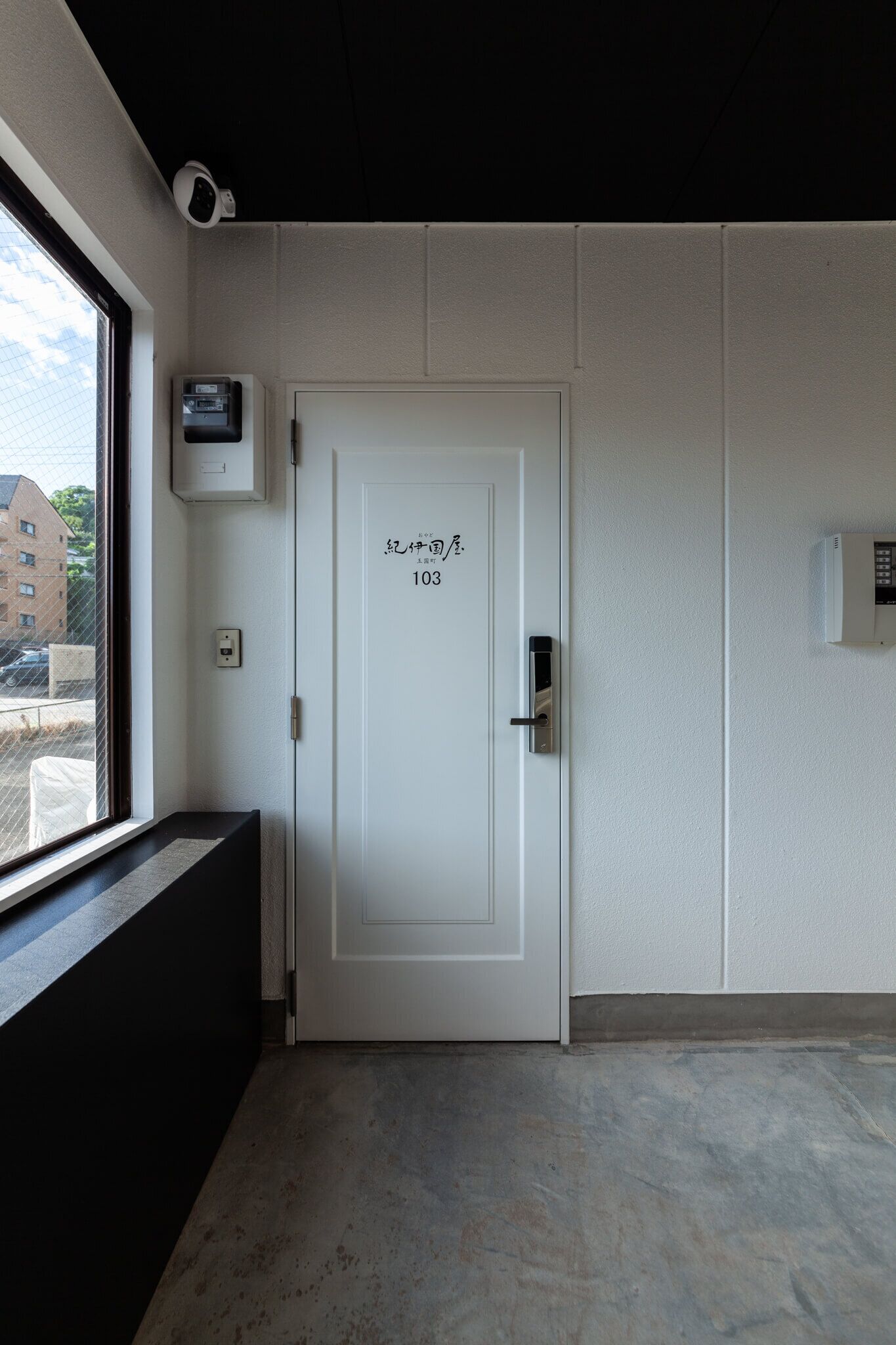 Habitación de hotel con una puerta blanca numerada, una ventana que ofrece vistas a edificios y una cámara de seguridad en la pared.