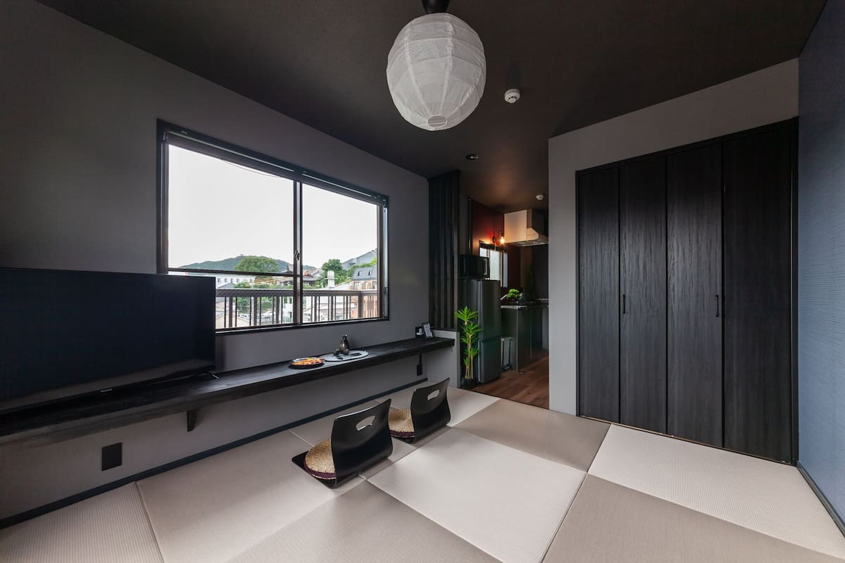 Una sala de estar moderna con un ventanal grande, un televisor de pantalla plana, esteras de tatami y un armario de madera oscura.