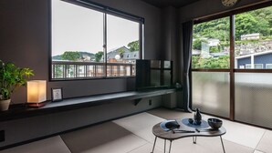 A modern room with tatami flooring, a television on a low shelf, a ceiling fan, and a large window offering a view of buildings and greenery. - Oyadokinokuniya tamazonomachi (Nagasaki)