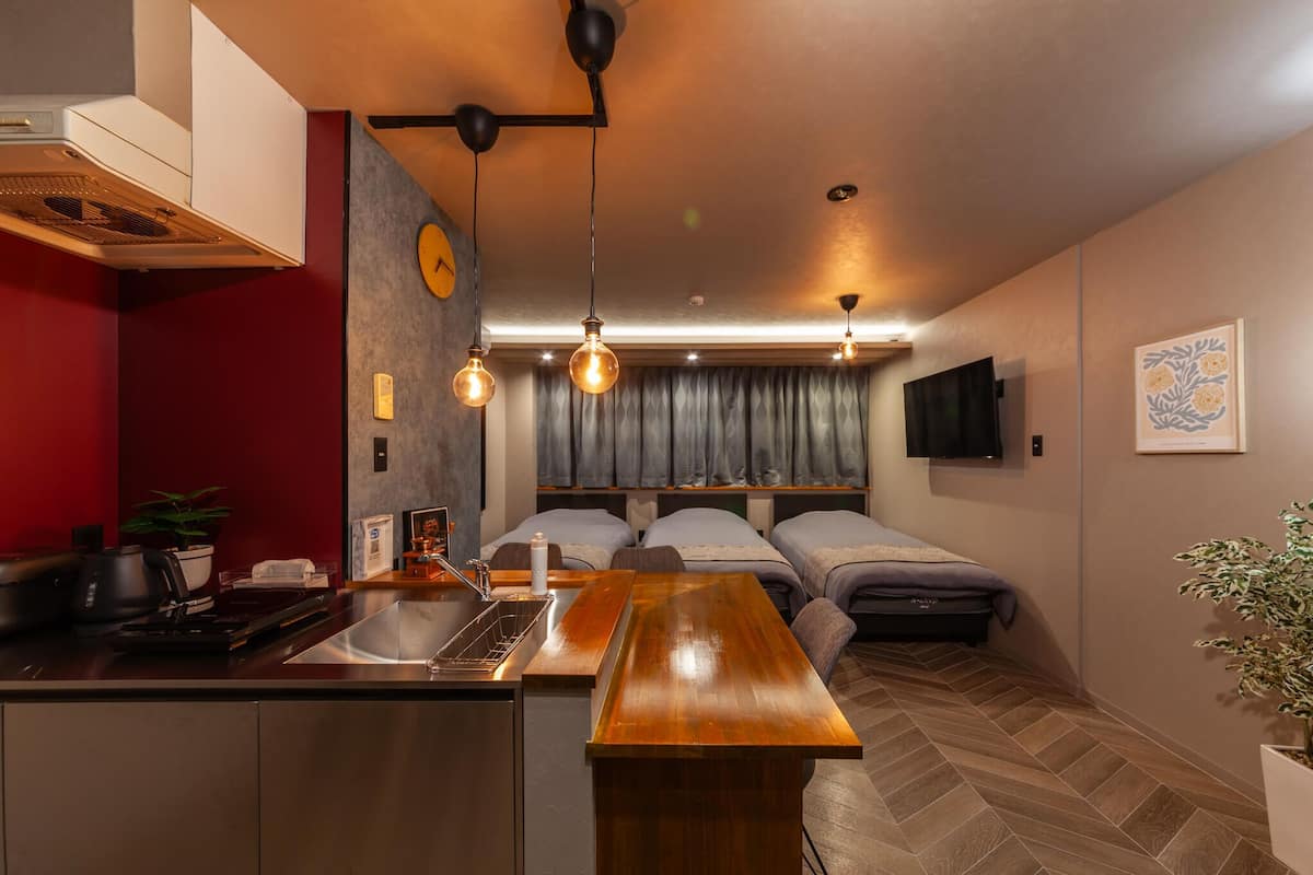 Habitación de hotel compacta con kitchenette, una mesa de comedor de madera, una cama con dos almohadas, un televisor montado en la pared y una planta en maceta.