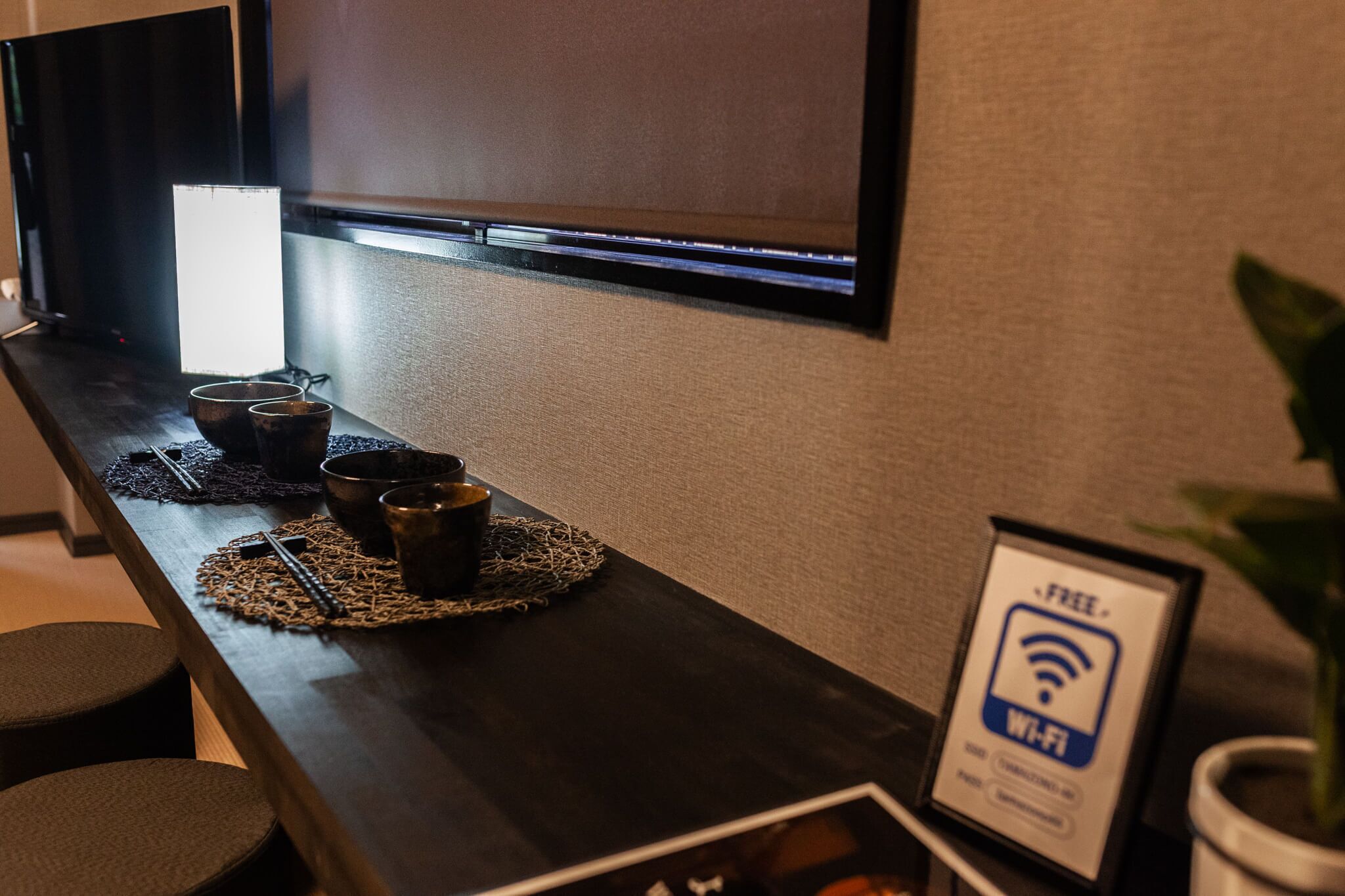 Un área de recepción de hotel con un mostrador de madera oscura, un televisor de pantalla plana montado en la pared, un letrero que indica Wi-Fi gratuito y una planta en maceta.