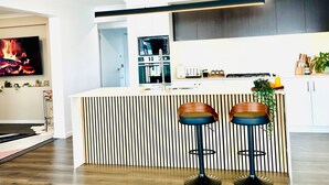 A modern kitchen with a central island, bar stools, and a flat-screen TV displaying a fireplace.