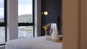 A bedroom with a large window, a bed with a textured blanket, a hanging pendant light, and a framed quote on the wall.