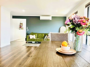 Un salon moderne avec un canapé vert, une table à manger sur laquelle se trouve un panier de fruits, et un vase rempli de fleurs.