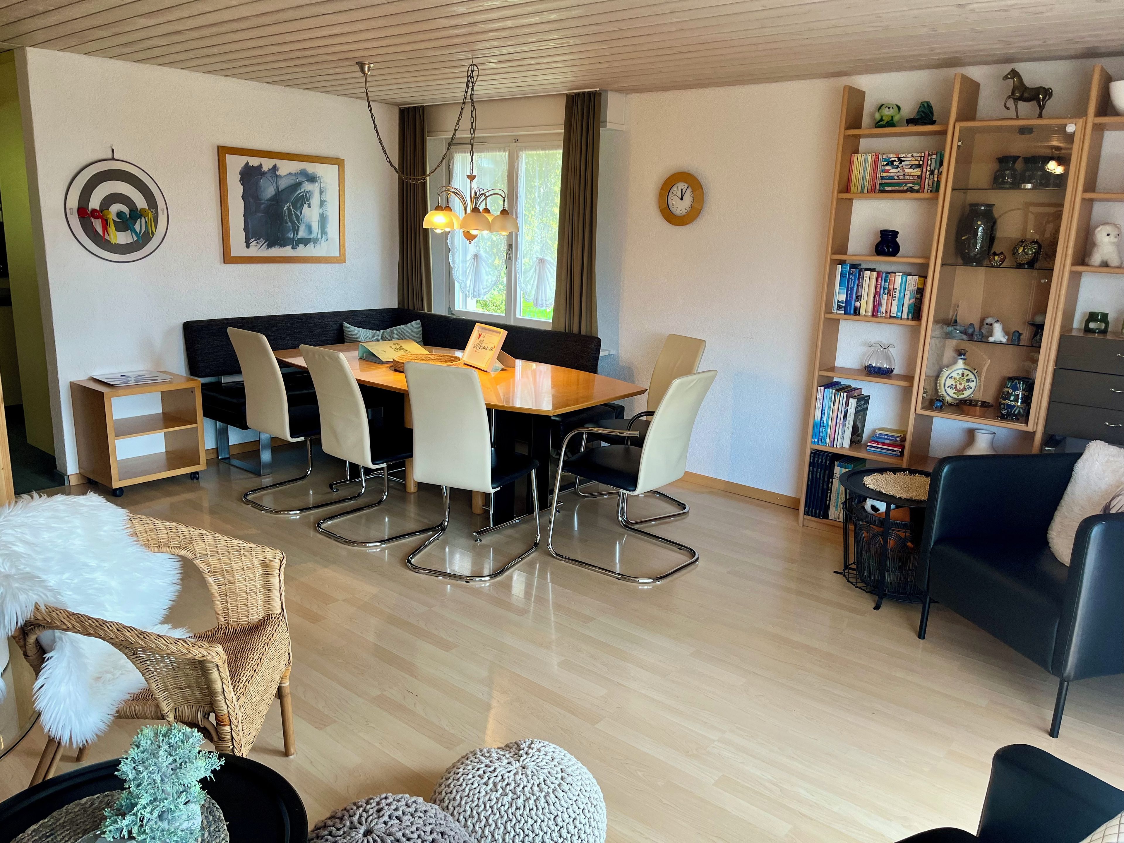 A modern living room with a dining area, featuring a wooden dining table, comfortable seating, a bookshelf, and a decorative wall clock.