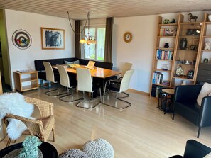 Una sala de estar moderna con comedor, que incluye una mesa de madera, asientos cómodos, una estantería y un reloj de pared decorativo.