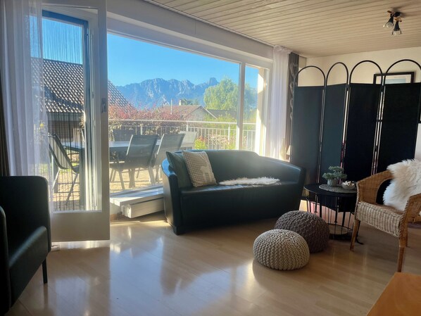 A modern living room with a large sliding glass door leading to an outdoor patio, a black sofa, a small round ottoman, a wicker chair, and a black metal partition.