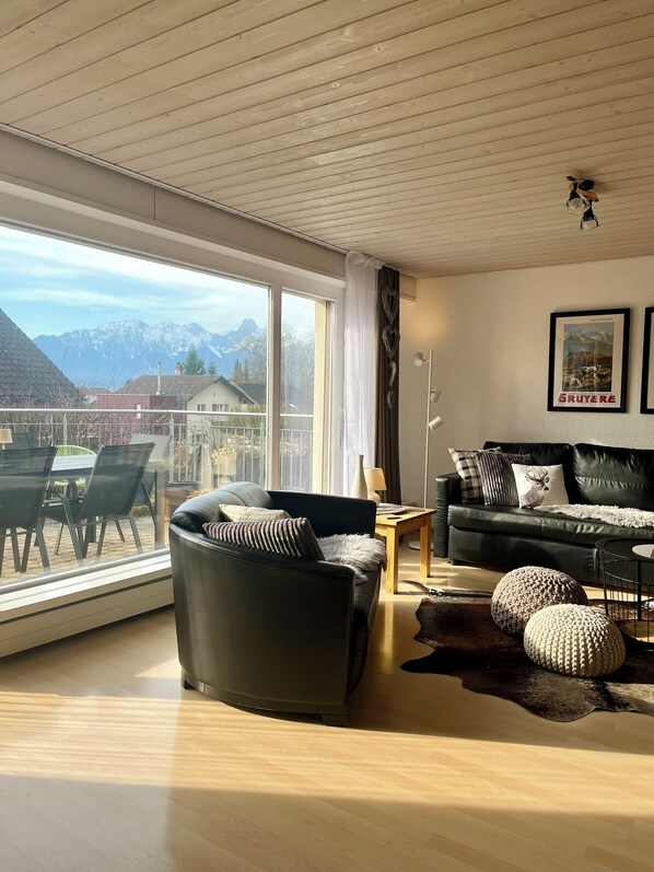 A modern living room with a large window, a leather sofa, a wooden coffee table, and a view of mountains.