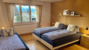 Un dormitorio con dos camas, una ventana con cortinas, un candelabro, una mesita de noche y una estantería con objetos.