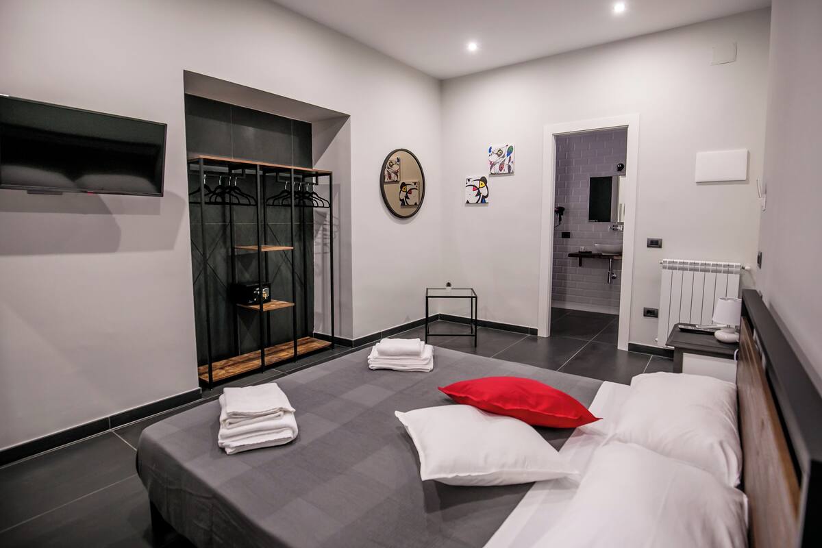 A modern hotel room with a bed, a small table, a mirror, and a bathroom visible through an open door.
