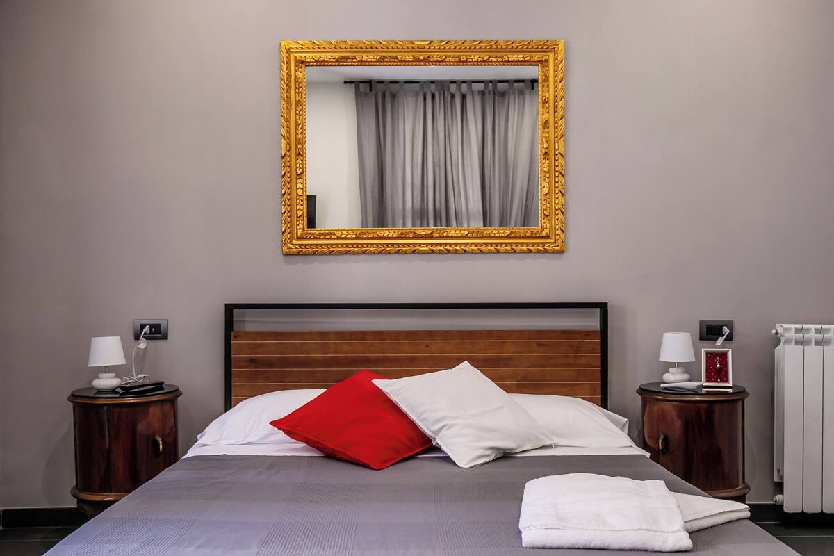 A neatly made bed with a grey checkered bedspread, a red pillow, and white pillows. There is a wooden headboard, a nightstand with a lamp, and a framed mirror on the wall.