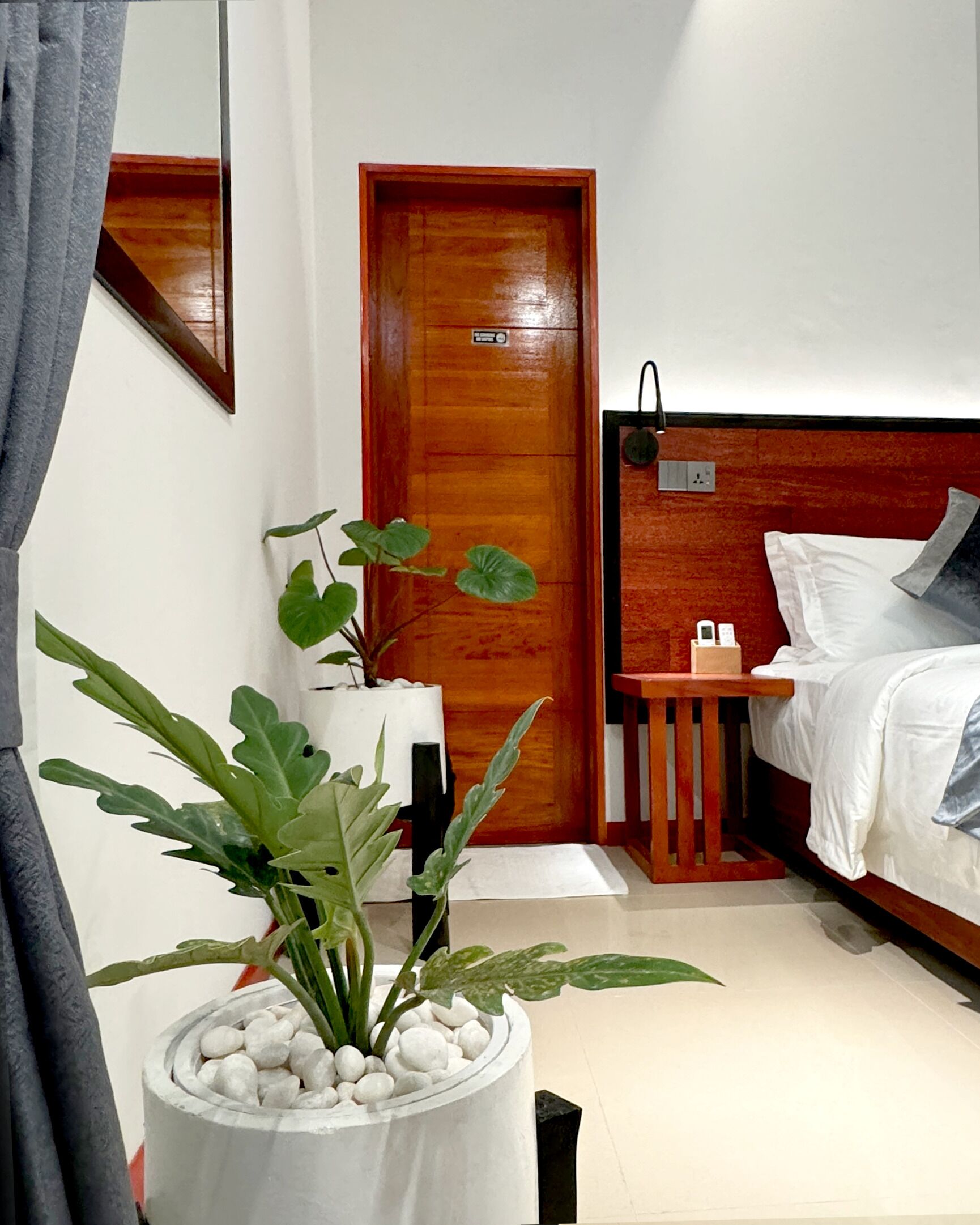 Quarto de hotel com porta de madeira, cama com roupa de cama branca, mesa de cabeceira com abajur e planta em vaso com pedrinhas brancas.