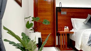 Una habitación de hotel con puerta de madera, cama con ropa de cama blanca, mesita de noche con lámpara y planta en maceta con guijarros blancos.