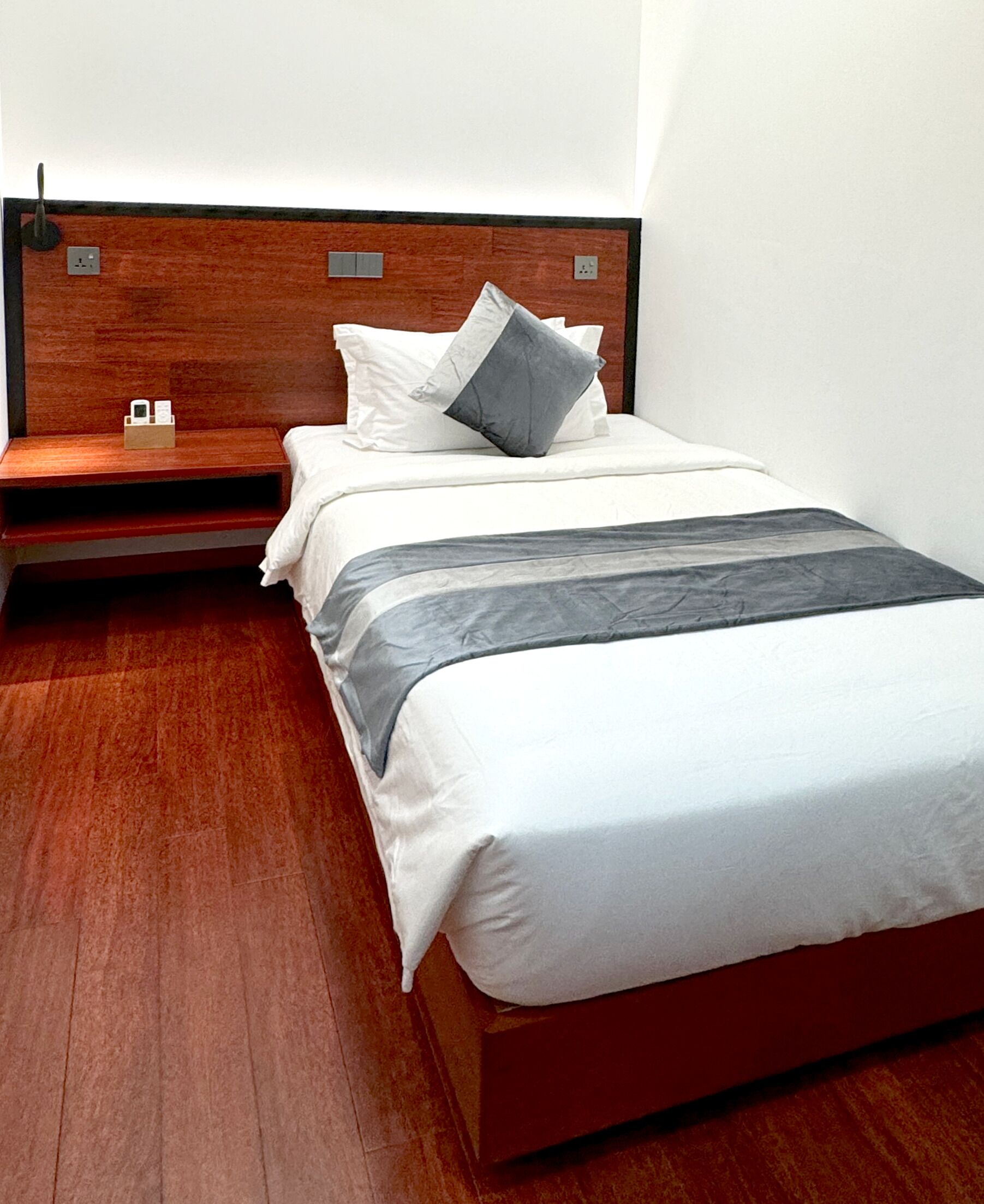 Ein modernes Hotelzimmer mit einem hölzernen Kopfteil, einem Bett mit weißen Leinen und einer grauen Tagesdecke sowie einem Nachttisch mit Telefon und einer kleinen Schachtel.