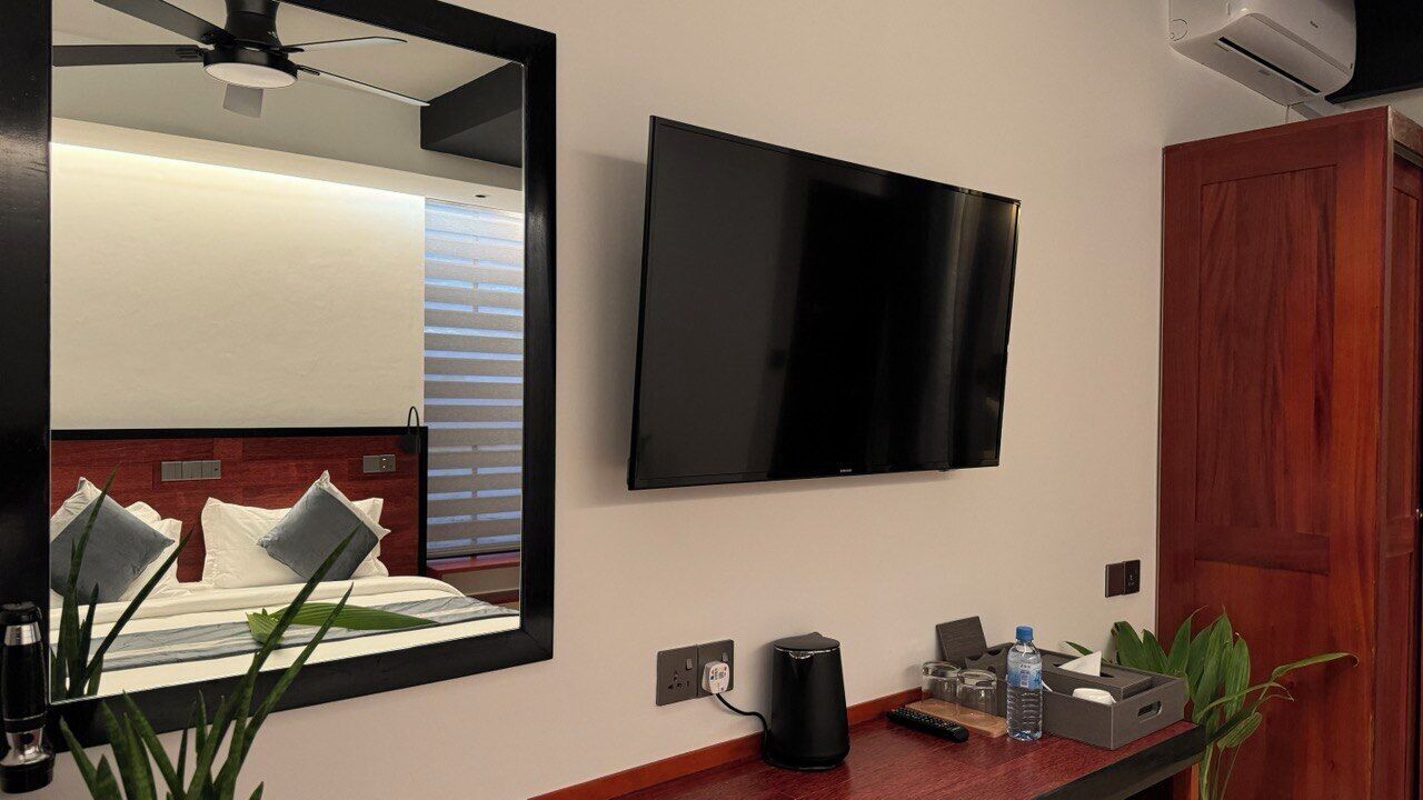 Habitación de hotel moderna con una amplia televisión de pantalla plana en la pared, una mesita de noche de madera con una tetera, una botella de agua y un control remoto.