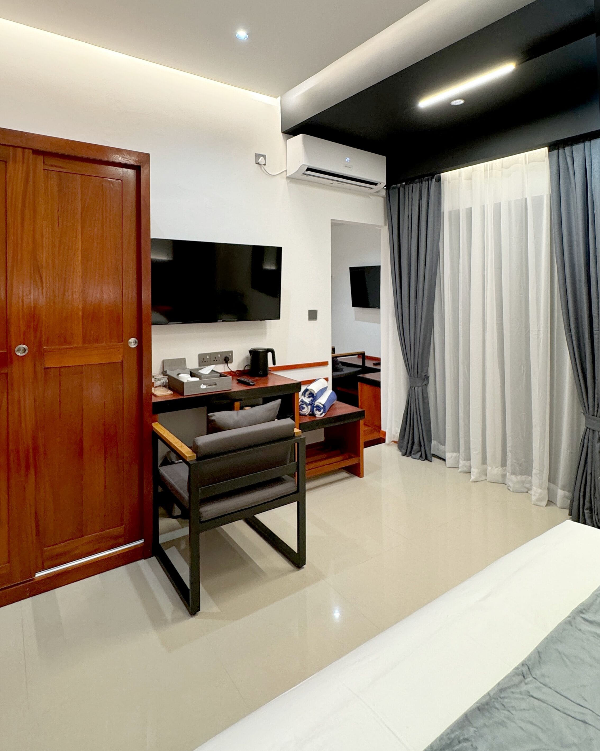 Een moderne hotelkamer met een houten deur, een flatscreen televisie aan de wand, een bureau met stoel, een bed en een groot raam met grijze gordijnen.