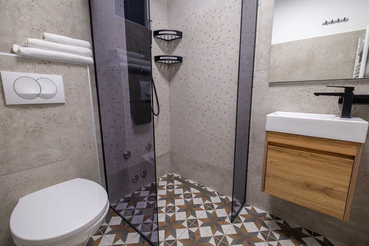 Un baño con inodoro, ducha con puerta de vidrio, lavamanos con gabinete de madera y piso de baldosas.