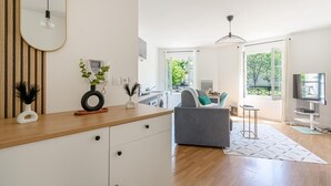 Een moderne woonruimte met een keukenhoek, een grijze bank, een televisie en een groot raam met uitzicht op de natuur.