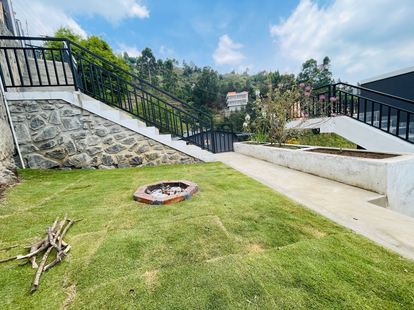 Una escalera de piedra que lleva a un edificio, una fogata en el patio, y una vista de una colina con árboles y una casa.