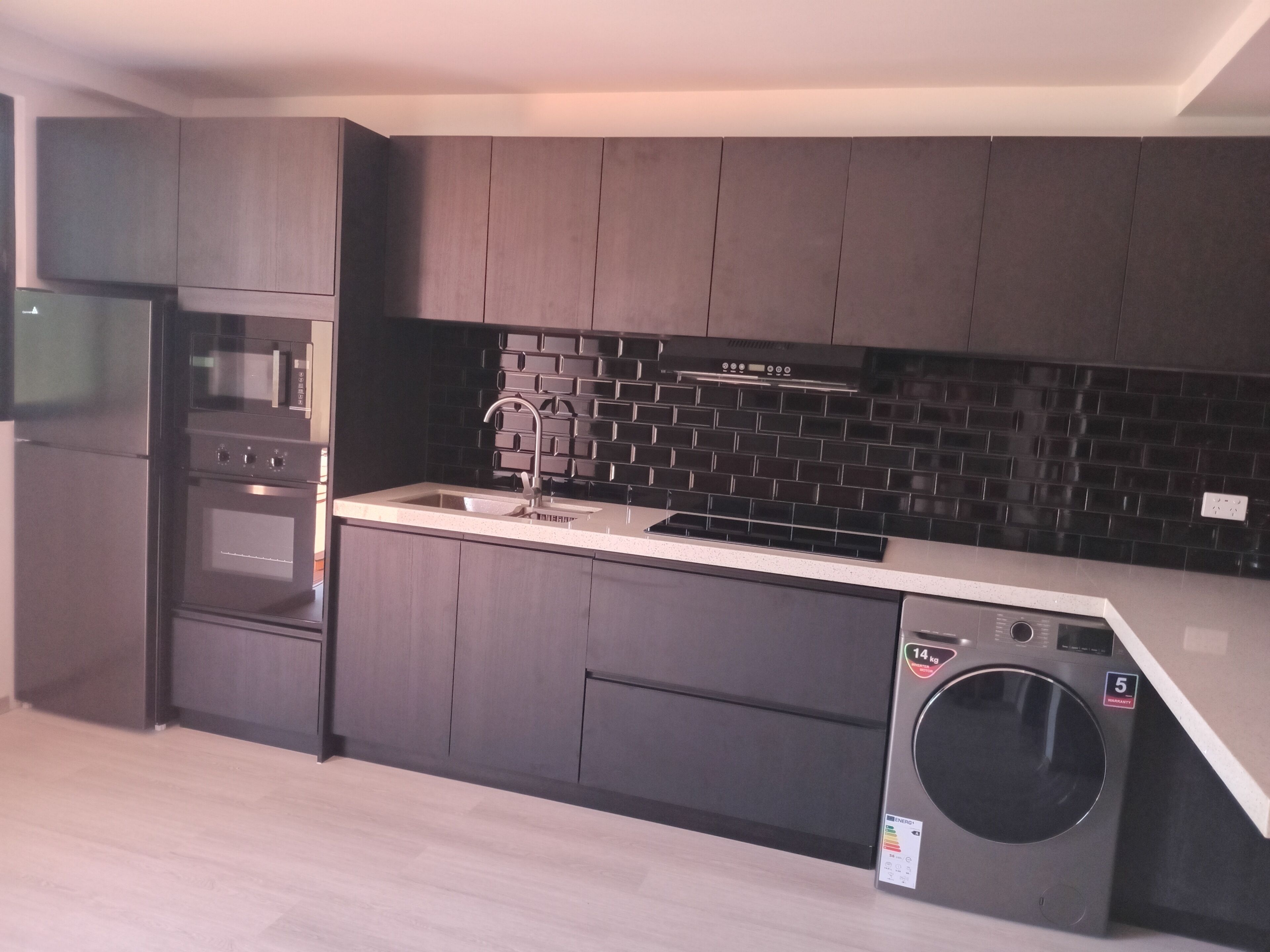 A modern kitchen with dark cabinetry, a stainless steel sink, a built-in oven, and a washing machine.