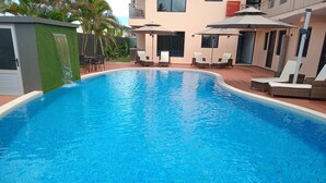 A swimming pool with lounge chairs and umbrellas, surrounded by a building with a balcony and a small waterfall feature. - Hibiscus Suites And Residences  (Nadi)