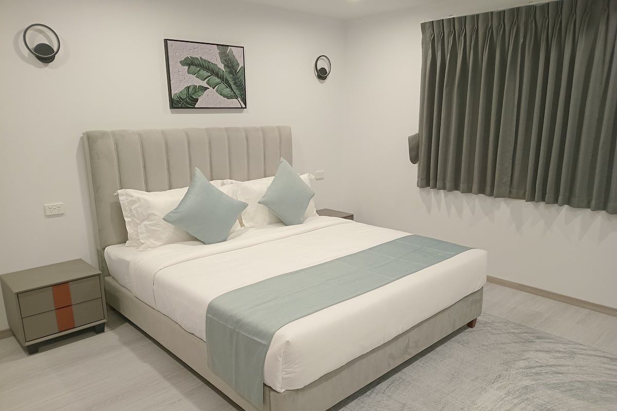 A bedroom with a bed, nightstands, a wall-mounted light, a window with curtains, and a framed picture on the wall.