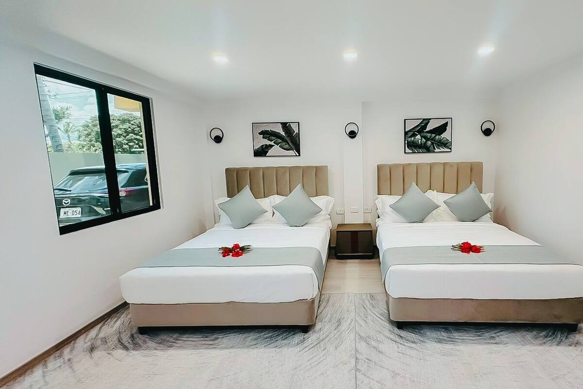 A hotel room with two beds, a window with a view of parked cars, and wall-mounted artwork.