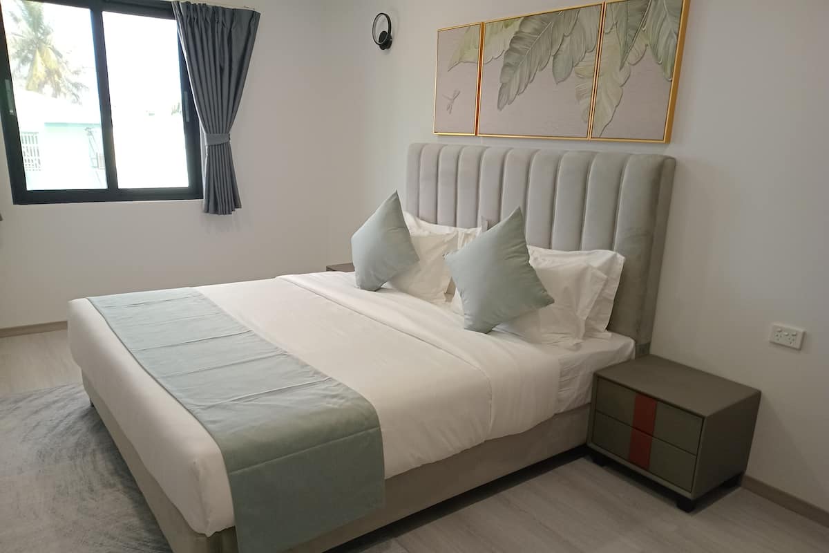 A bedroom with a bed, a nightstand, a wall-mounted light, a window with curtains, and a framed leaf artwork.