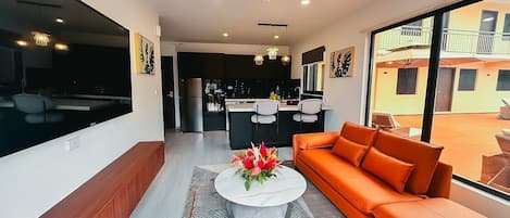 Una sala de estar moderna con un sofá, una mesa de centro y un televisor. Se ve un área de cocina al fondo, con un refrigerador y una mesa de comedor.