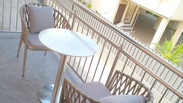 Un balcón con una mesa y dos sillas, baranda metálica y un edificio con varias ventanas y unidades de aire acondicionado al fondo.