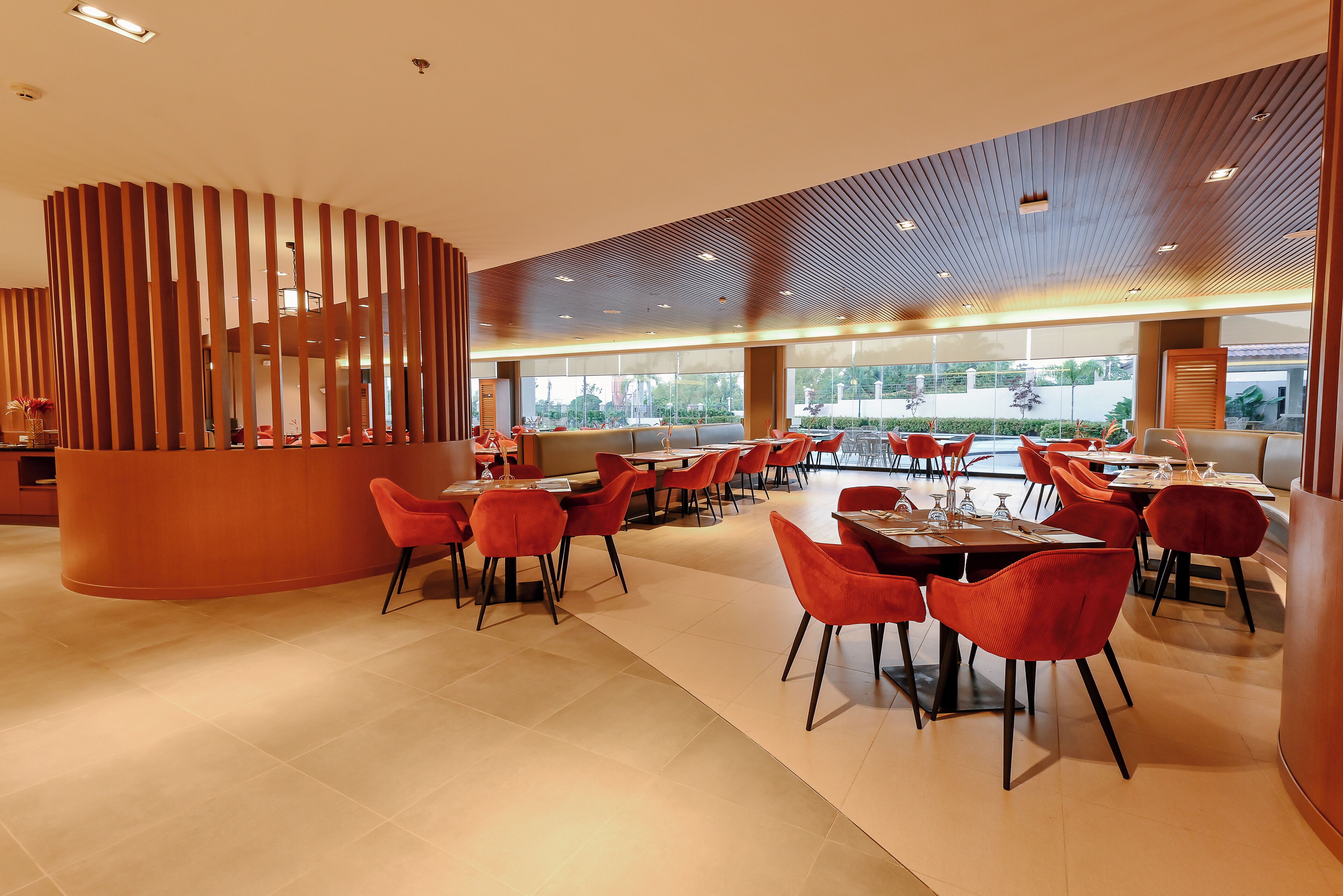 A modern restaurant with a curved wooden partition, red chairs, and a spacious layout with large windows.