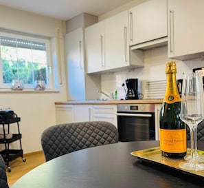 A modern kitchen with white cabinets, a dining table with a bottle of champagne and a glass, and a window with a view of greenery. - Antonia Vacation Home (Varel)