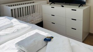 Ein Hotelzimmer mit Reisebett, Fernseher auf einer Kommode und ordentlich gefalteten Handtüchern auf dem Bett.