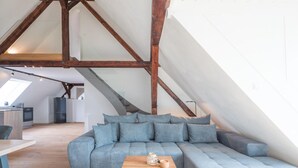 Une chambre à coucher de style loft moderne, avec des poutres en bois apparentes, un grand canapé d’angle gris, une table basse en bois et une lucarne.