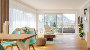 A spacious living area with a wooden dining table, teal chairs, a sofa, and a large sliding glass door leading to a balcony with a mountain view.