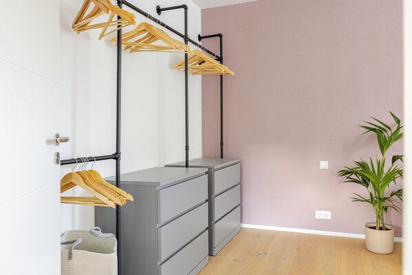 Una habitación con un aparador gris, perchas de madera en un soporte de metal negro, una planta en maceta y una pared blanca.