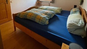Un lit simple avec un matelas bleu et une couette à carreaux, un plafond en bois, une fenêtre avec des rideaux et un radiateur.