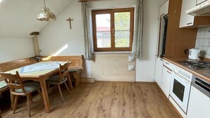 Une cuisine avec un plancher en bois, une table à manger avec des chaises, une fenêtre avec des rideaux et un poêle.
