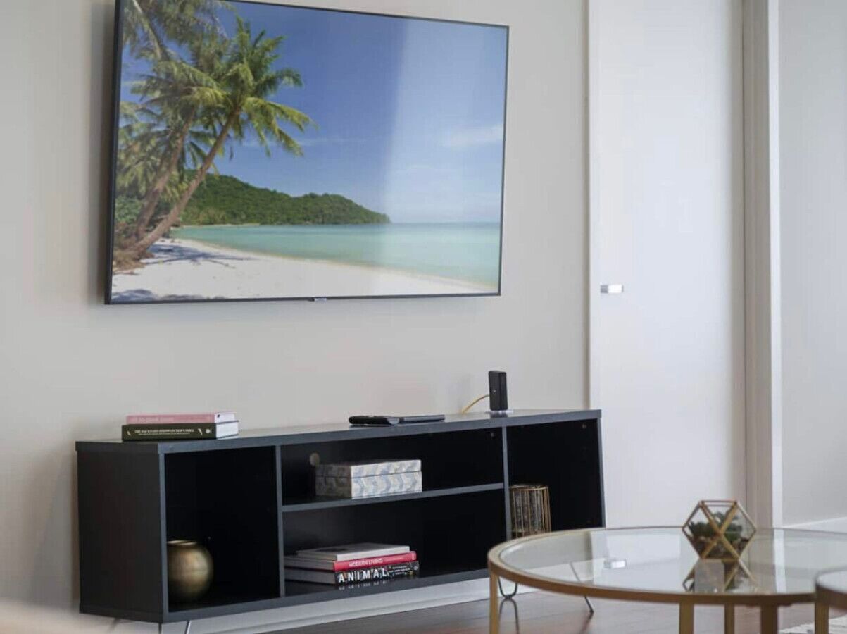 A living room with a flat-screen TV mounted on the wall, a black entertainment unit with books and a speaker, and a round glass coffee table.