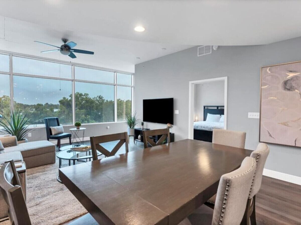 A spacious living room with a large dining table, comfortable seating, a ceiling fan, and a large window offering a view of the outdoors.
