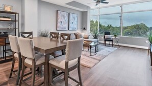 A modern dining area with a wooden table and chairs, a sofa, and a view of the outdoors through large windows. - Luxury 1260sq ft 2 Bd/2BTH in DT (Orlando)