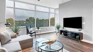 A modern living room with a large window, a ceiling fan, a flat-screen TV, and a minimalist furniture arrangement. - Luxury 1260sq ft 2 Bd/2BTH in DT (Orlando)
