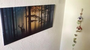Una habitación con una pared blanca texturizada, un cuadro de un paisaje boscoso, una pequeña silla de madera y una guirnalda de adornos con forma de casitas para pájaros.