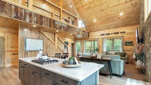 Una cabaña espaciosa con interior de madera, una isla central con electrodomésticos de cocina, un área de estar con sofá y sillones, y una escalera que conecta los niveles.