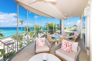 A balcony with a view of the ocean and palm trees, furnished with wicker chairs and a table. - The Meridian Villa 25, 3BR/3B Oceanfront Fourth Floor, fully equipped. (Seven Mile Beach)