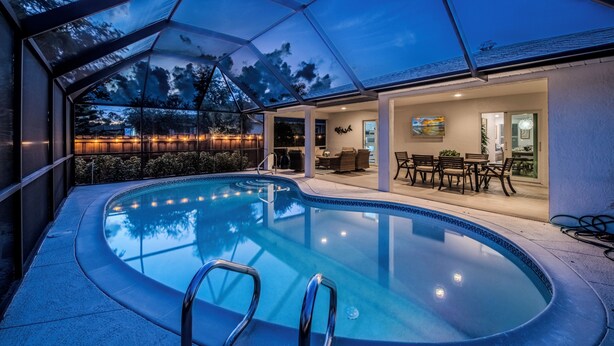 A modern indoor pool with glass walls, a lounge area with seating, and a view of the outdoors. - Pet-Friendly 3-Bedroom Home with Pool (Bonita Springs)