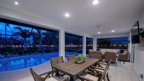 A modern outdoor dining area with a wooden table and chairs, a pool, and a view of the surrounding landscape. - Pet-Friendly 3-Bedroom Home with Pool (Bonita Springs)